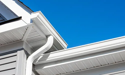 A close-up of a new white gutter, downspout, and vented soffit system on a grey-sided home, installed by Silverline Roofing Ltd against a clear blue sky